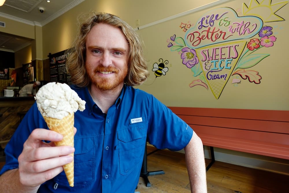 Samuel Jeffery holding an ice cream cone in front of a colourful hand-painted mural inside a shop at Heritage Park in Calgary, Alberta, Canada, during summer.