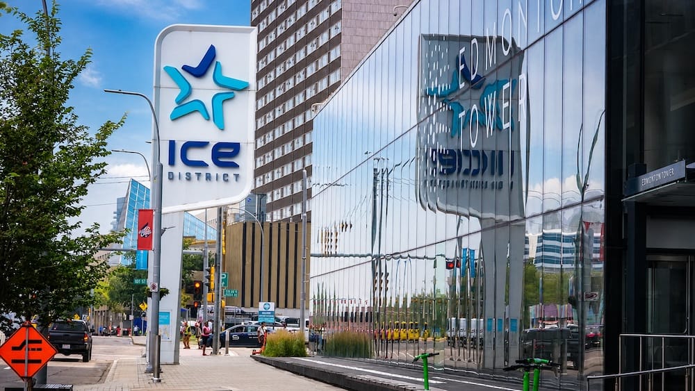 El cartel del ICE District en una esquina del centro de Edmonton junto a la fachada de vidrio espejado de la Edmonton Tower, que refleja su propio cartel de "Edmonton Tower", bajo un cielo despejado de verano con peatones y tráfico abajo.
