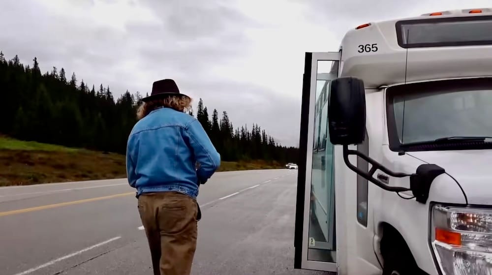 Samuel Jeffery camina hacia una van blanca de excursión estacionada junto a la Icefields Parkway en Alberta, Canadá, con la puerta abierta, la ruta y laderas cubiertas de pinos alrededor.