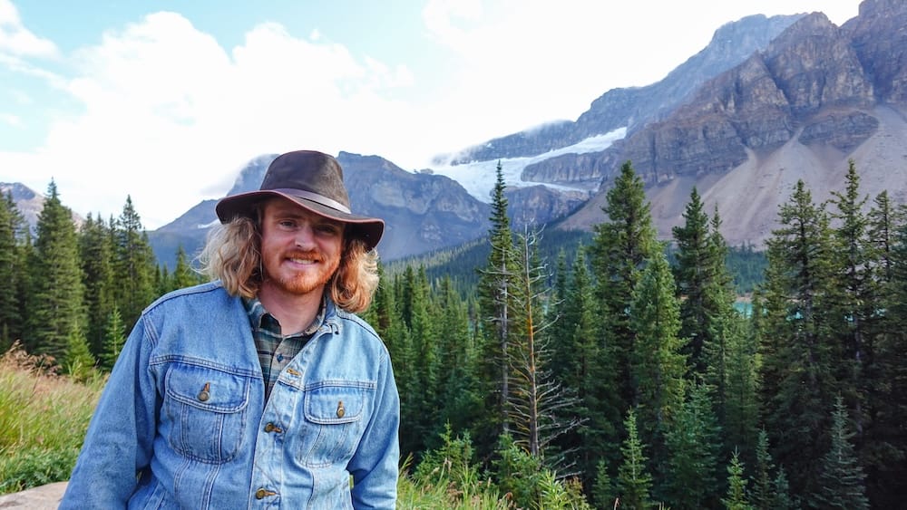 Samuel Jeffery está junto a un bosque de pinos en la Icefields Parkway, Alberta, Canadá, con acantilados montañosos y un glaciar visibles bajo un cielo que empieza a despejarse.