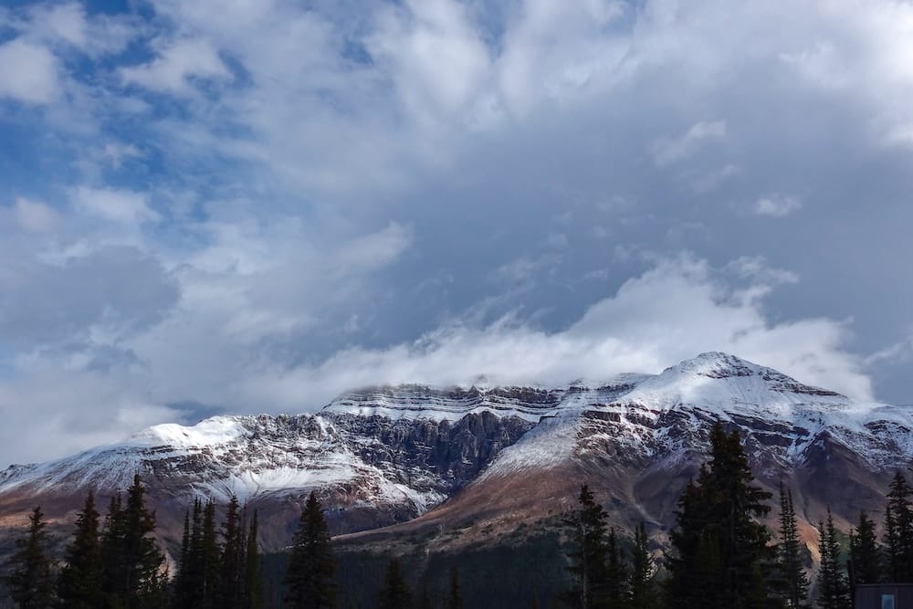Picos de las Montañas Rocosas cubiertos parcialmente de nieve se elevan sobre un bosque oscuro de pinos junto a la Icefields Parkway, Alberta, Canadá, bajo nubes y sectores de cielo azul.