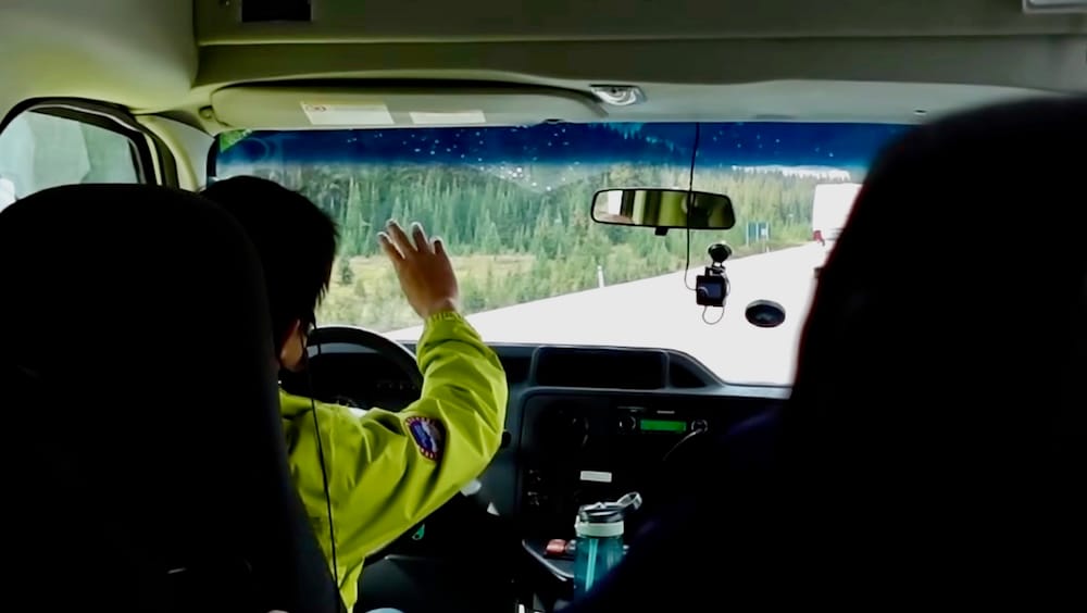 Combi de tour guiado en la Icefields Parkway — Parque Nacional Banff, Alberta, Canadá Vista desde adentro de una combi de tour de grupo reducido en la Icefields Parkway, el guía señalando la ruta de montaña boscosa más adelante bajo cielos despejados, Parque Nacional Banff, Alberta, Canadá.