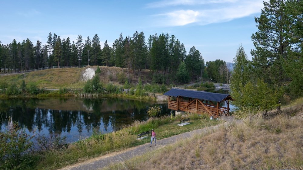 Idlewild Park in Cranbrook, British Columbia, seen from an elevated viewpoint showing the full lake loop trail, calm reflective water, pine forest hillsides, and benches that make this peaceful park ideal for relaxed walks and scenery.
