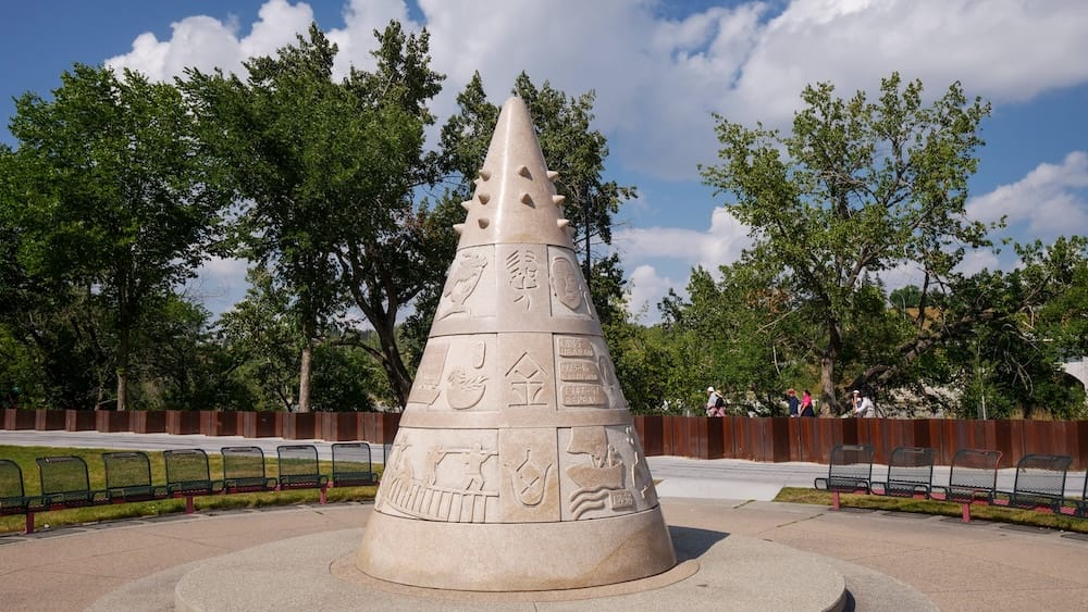 The conical granite sculpture "In Search of Gold Mountain" carved with layered relief scenes and Chinese characters, standing in a seating area at Sien Lok Park in Calgary, Alberta, Canada.
