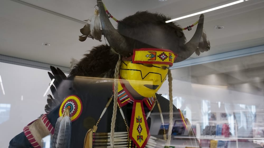 Una vitrina de ropa ceremonial indígena que incluye un tocado con cuernos, escarapelas con mostacillas y una corbata de mostacillas, parte de una exhibición de arte dentro de la Biblioteca Central de Calgary en Calgary, Alberta, Canadá.