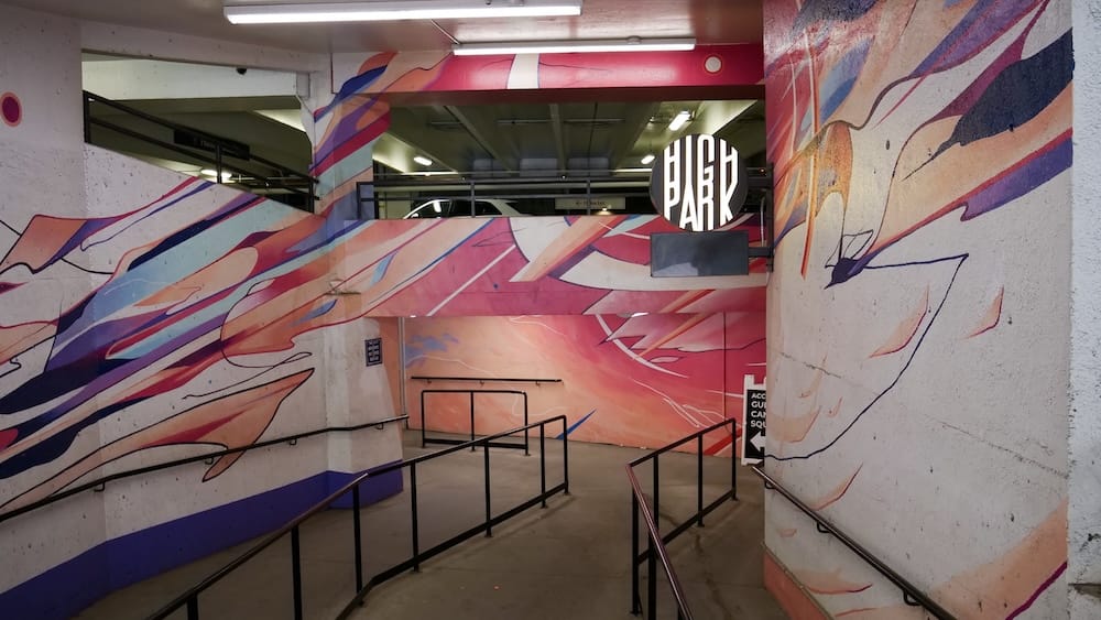 Un amplio mural abstracto en tonos rosa, coral y azul cubre las paredes de la escalera y el pasillo que lleva a High Park, con un cartel circular negro de "High Park" visible más adelante cerca del interior del estacionamiento.