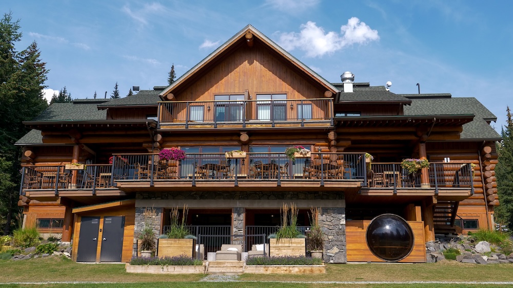 Island Lake Lodge accommodations in Fernie, British Columbia, showing the timber-and-stone main lodge with balconies, flower boxes, and mountain lodge architecture set against forest surroundings, highlighting the upscale yet rustic lodging available for overnight guests.
