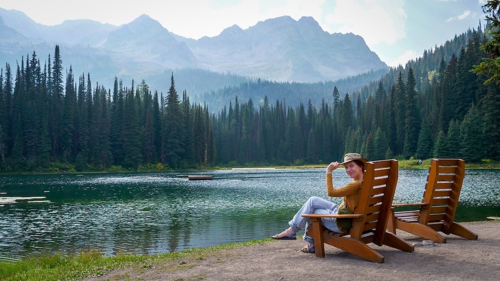 Island Lake Lodge near Fernie, British Columbia, showcasing tranquil alpine lake scenery as Audrey Bergner relaxes in a wooden chair by the water, surrounded by evergreen forest and rugged Rocky Mountain peaks on one of the most iconic and peaceful day trips from Fernie.