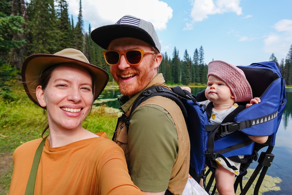 Island Lake Lodge hike in Fernie, British Columbia, with Nomadic Samuel Jeffery and Audrey Bergner of That Backpacker smiling on the lakeside trail while carrying their baby Aurelia in a backpack carrier, surrounded by alpine forest and calm mountain water.
