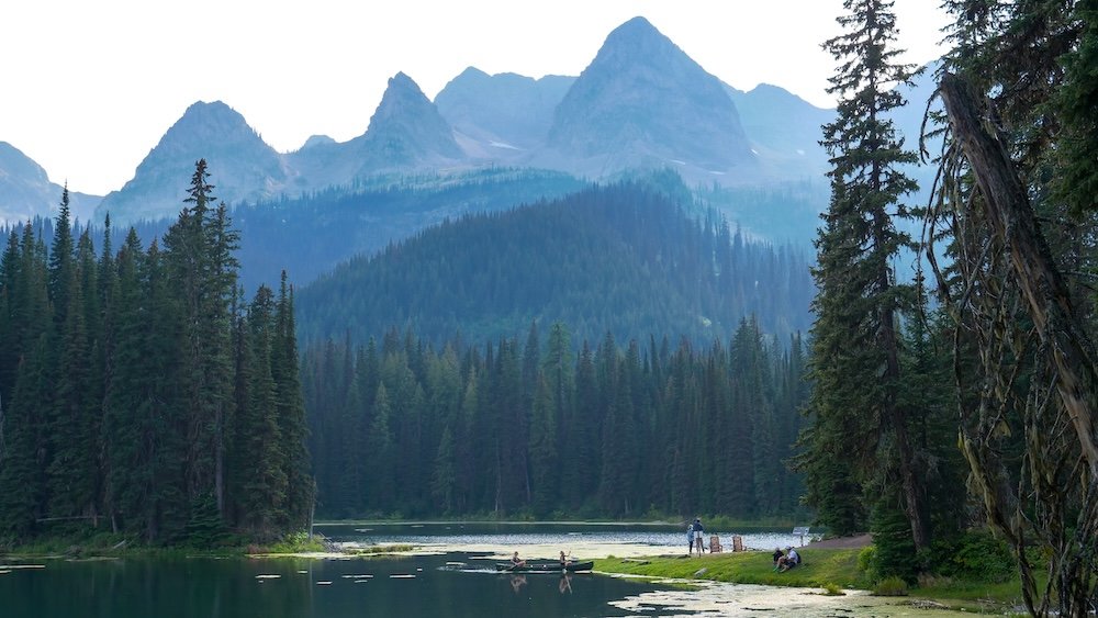 Canoeing at Island Lake Lodge, Fernie Island Lake Lodge in Fernie, BC shows a calm alpine lake with canoes on the water, surrounded by dense evergreen forest and dramatic Rocky Mountain peaks rising in the background on a peaceful summer day.