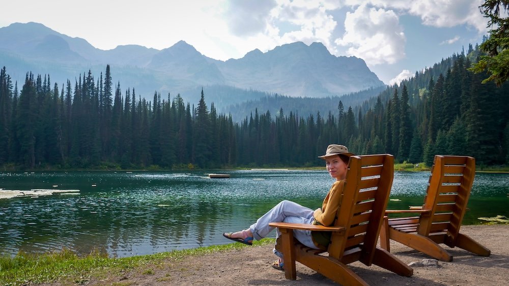 Island Lake Lodge lakeside chairs and mountain views Island Lake Lodge in Fernie, BC: Audrey Bergner of That Backpacker relaxes in a wooden lakeside chair, looking across emerald water toward dense pine forest and rugged Rocky Mountain peaks on a calm summer afternoon.