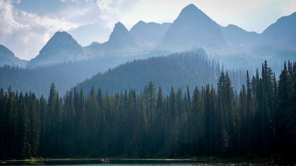 Tiny canoe on Island Lake beneath Fernie’s mountain peaks A tiny canoe glides across the still waters of Island Lake Lodge in Fernie, British Columbia, framed by dense evergreen forest and dramatic, jagged mountain peaks rising in hazy layers behind it, creating a serene and imposing backcountry landscape.