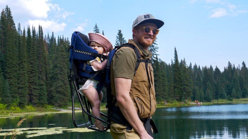 Island Lake Lodge lakeside hike with baby carrier Island Lake Lodge in Fernie, BC: Nomadic Samuel Jeffery hikes beside the lake with baby Aurelia in a backpack carrier, passing a pine-lined shoreline with calm water and mountain views on a bright summer afternoon.