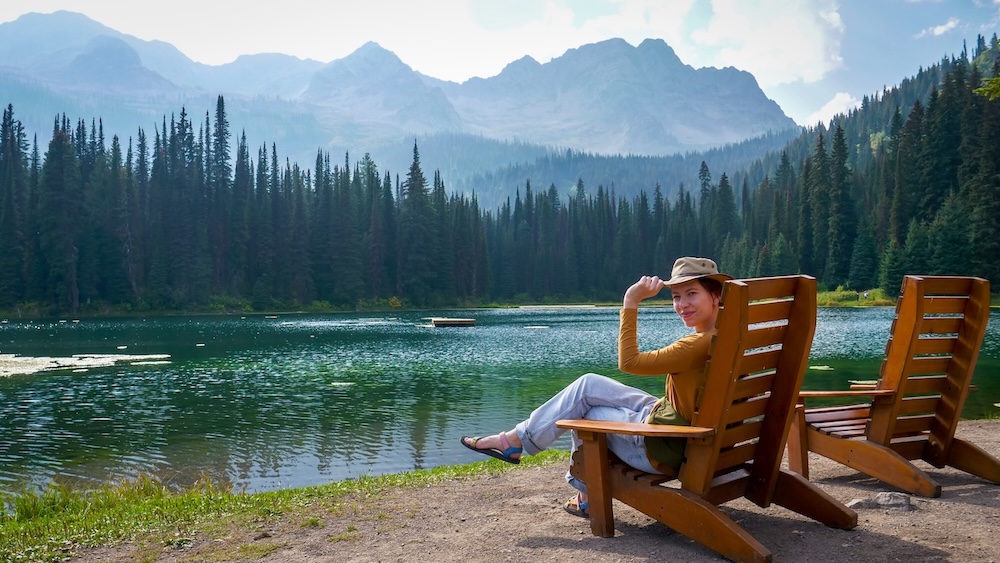 Island Lake Lodge in Fernie, British Columbia, with calm alpine lake, wooden chairs on the shore, and forested peaks rising behind, as Audrey Bergner relaxes by the water enjoying a quiet pause after lunch and an easy lakeside walk.
