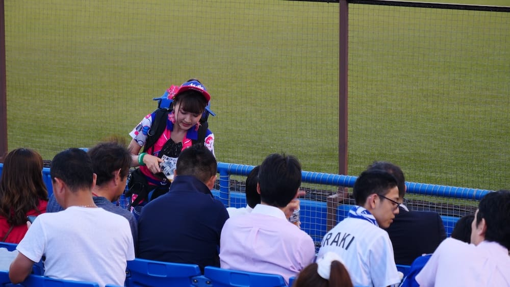 A Japanese beer vendor serves spectators from the aisle beside the outfield seats during an NPB baseball game at Meiji Jingu Stadium in Tokyo, Japan, with fans seated nearby.
