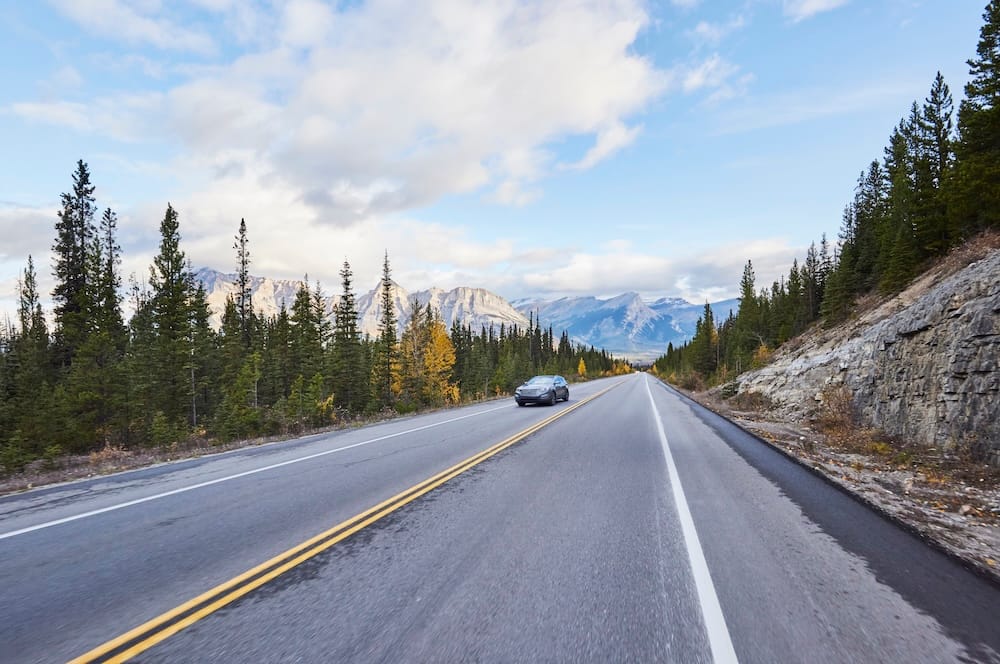 A car travelling along a mountain highway in Jasper National Park, Alberta, Canada, with evergreen forest, exposed rock and the Canadian Rockies stretching across the horizon.