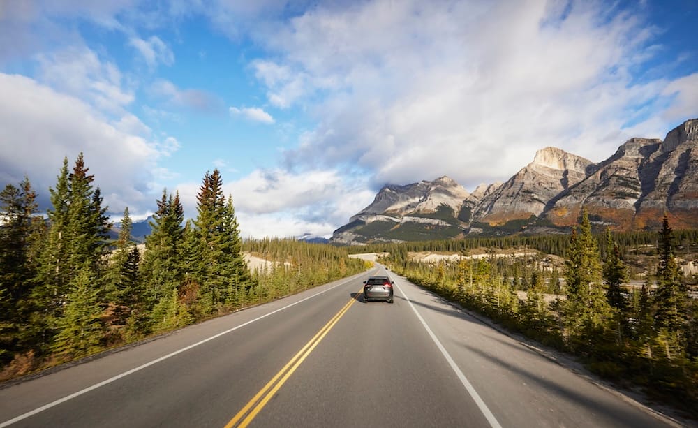 Scenic highway through Jasper National Park in Alberta, Canada, with a car travelling between evergreen forest and rugged Canadian Rockies beneath a partly cloudy blue sky.