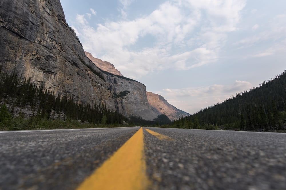 A low-angle view along a highway in Jasper National Park, Alberta, Canada, with yellow centre lines leading between steep Rocky Mountain cliffs and dense evergreen forest.