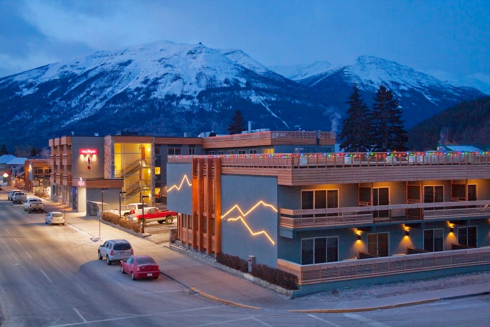 Jasper town in Alberta, Canada, with low-rise buildings and parked cars beneath snow-covered Canadian Rockies, photographed in the evening with warm lights glowing against the blue mountain backdrop.
