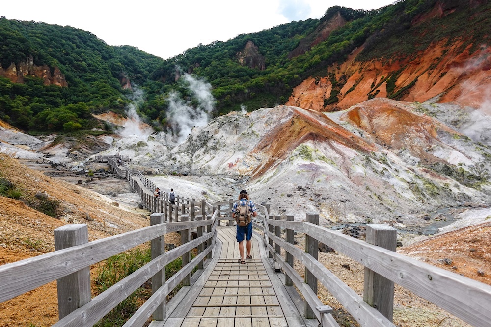 Samuel Jeffery walks along a winding wooden boardwalk deep into Jigokudani (Hell Valley), toward steam plumes and orange, white and grey volcanic slopes in Noboribetsu Onsen, Hokkaido, Japan.