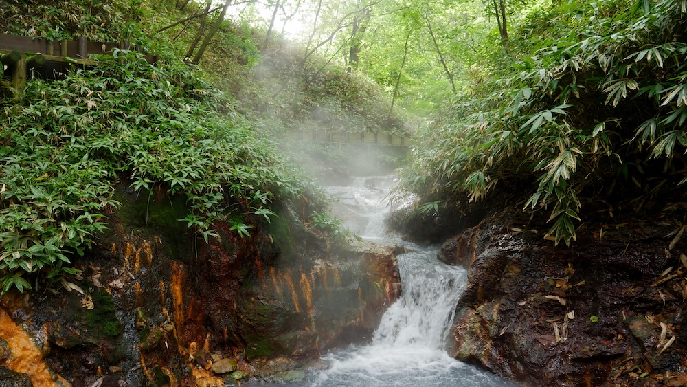Steaming Hot-Spring Cascade and Forest Mist on the Oyunuma River, Natural Footbath, Noboribetsu Onsen, Hokkaido, Japan Steam drifts through the forest above a small cascade of hot-spring water tumbling past orange-streaked rocks and bamboo grass on the Oyunuma River at the natural footbath in Noboribetsu Onsen, Hokkaido, Japan.