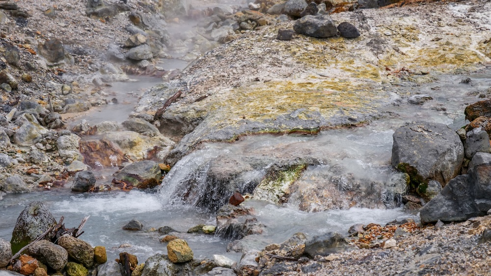 Geothermal Hot Water Flowing Over Mineral-Stained Rocks at Jigokudani Hell Valley – Noboribetsu Onsen, Hokkaido, Japan Close-up of steaming geothermal water cascading over grey rocks into a milky-blue stream at Jigokudani Hell Valley in Noboribetsu Onsen, Hokkaido, Japan, with yellow mineral-stained ground and scattered volcanic stones.