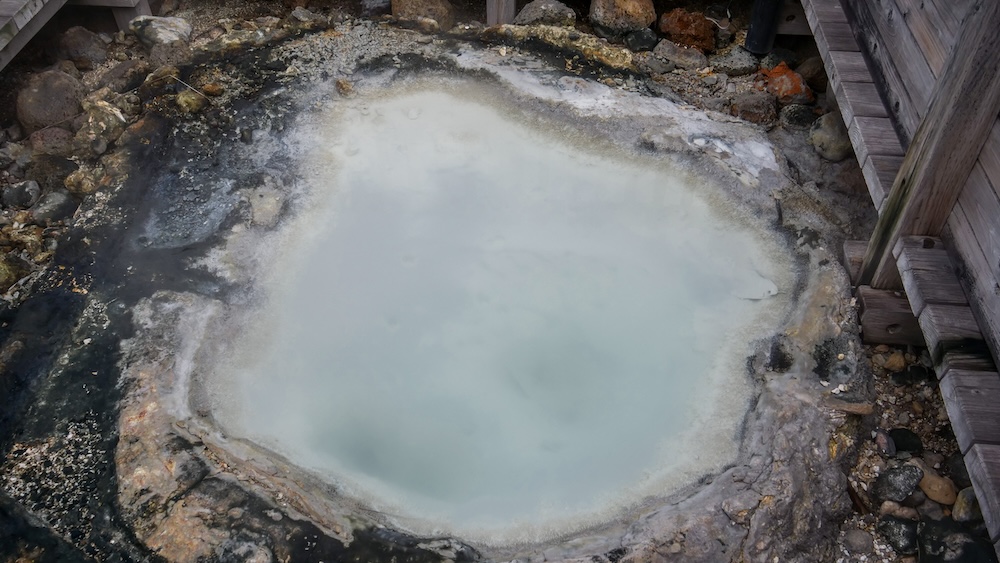 Milky-Blue Geothermal Pool With Mineral-Crusted Edges at Jigokudani Hell Valley – Noboribetsu Onsen, Hokkaido, Japan Close-up of a milky pale-blue geothermal pool at Jigokudani Hell Valley in Noboribetsu Onsen, Hokkaido, Japan, surrounded by dark volcanic rock, grey mineral deposits and wooden boardwalk structures along its edges.