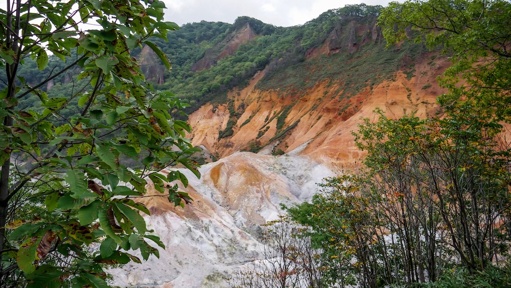 Jigokudani, Valle del Infierno de Noboribetsu Onsen, Hokkaido, Japón: laderas volcánicas anaranjadas enmarcadas por el bosque Laderas volcánicas anaranjadas y blancas cubiertas de minerales en Jigokudani, el Valle del Infierno de Noboribetsu Onsen, Hokkaido, Japón, con pequeñas fumarolas entre cerros rocosos enmarcados por árboles y arbustos verdes.