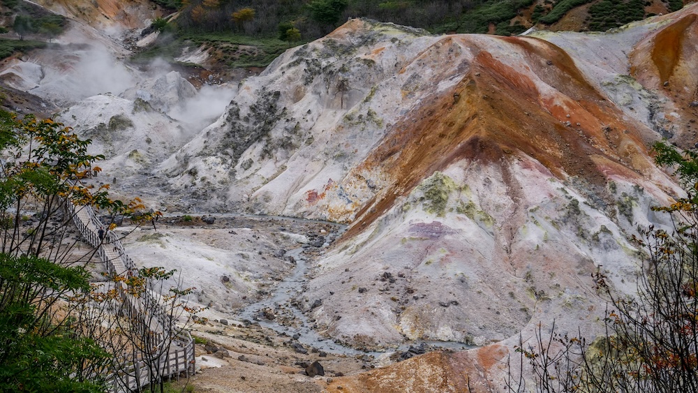 Colourful Volcanic Ridges and Winding Boardwalk at Jigokudani Hell Valley – Noboribetsu Onsen, Hokkaido, Japan Orange, rust-red and pale white volcanic ridges dominate Jigokudani Hell Valley in Noboribetsu Onsen, Hokkaido, Japan, with steam rising from the rocky basin and a wooden boardwalk winding through the lower-left foreground.