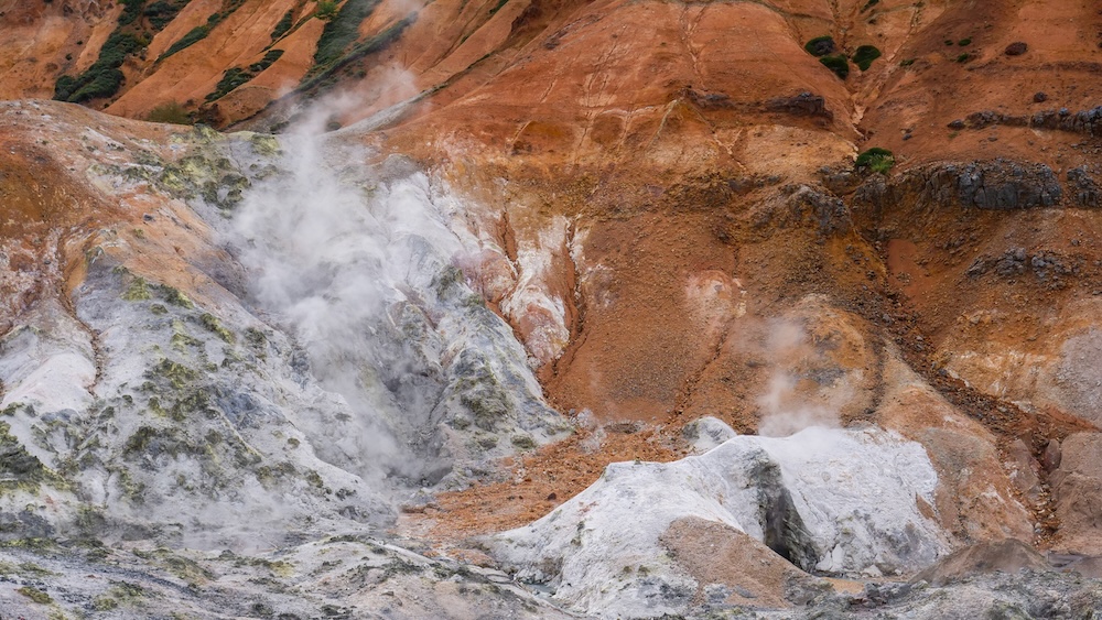 Fumarolas humeantes y laderas volcánicas teñidas de minerales en Jigokudani, Noboribetsu Onsen, Hokkaido, Japón Vapor geotérmico blanco sale de fumarolas entre rocas pálidas cubiertas de minerales y laderas volcánicas de color naranja oxidado en Jigokudani, el Valle del Infierno de Noboribetsu Onsen, Hokkaido, Japón, con pequeños sectores de vegetación verde.