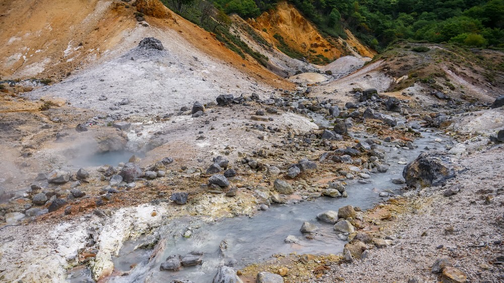 A steaming geothermal stream winding between grey and orange mineral-stained volcanic slopes at Jigokudani Hell Valley in Noboribetsu Onsen, Hokkaido, Japan, with scattered dark rocks, bubbling water and green forest in the background.