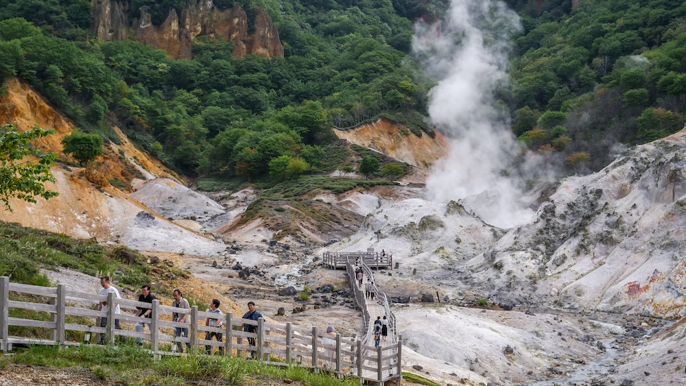 Jigokudani, el Valle del Infierno de Noboribetsu Onsen, Hokkaido, Japón: fumarolas y pasarelas de madera entre laderas volcánicas Pasarelas de madera con visitantes atravesando Jigokudani, el Valle del Infierno de Noboribetsu Onsen, Hokkaido, Japón, mientras una gran columna de vapor blanco se eleva desde laderas volcánicas claras rodeadas de bosque.