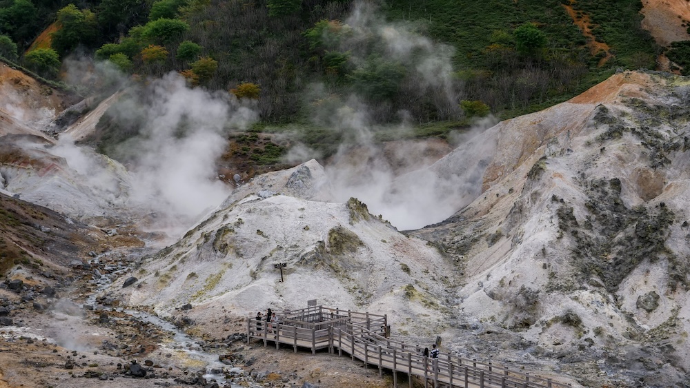 Paisaje volcánico de Jigokudani, Valle del Infierno, y pasarela de observación en Noboribetsu Onsen, Hokkaido, Japón Vista panorámica de Jigokudani, el Valle del Infierno, en Noboribetsu Onsen, Hokkaido, Japón. Grandes columnas de vapor geotérmico salen de laderas volcánicas claras, teñidas por minerales, mientras varios visitantes recorren una pasarela de madera.