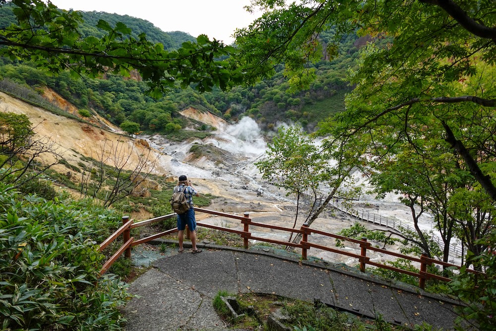Samuel Jeffery fotografiando Jigokudani desde un mirador entre árboles en Noboribetsu Onsen, Hokkaido, Japón Samuel Jeffery fotografía las laderas volcánicas humeantes de Jigokudani, el Valle del Infierno de Noboribetsu Onsen, Hokkaido, Japón, desde un mirador rodeado de árboles, con barandas rojas, una pasarela sinuosa y colinas verdes.