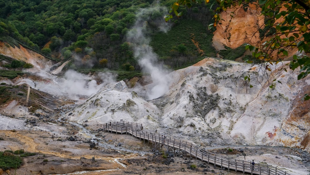 Pasarela de madera que atraviesa el paisaje volcánico de Jigokudani, el Valle del Infierno, en Noboribetsu Onsen, Hokkaido, Japón, con vapor blanco saliendo de fumarolas, laderas rocosas claras manchadas de minerales y cerros boscosos.