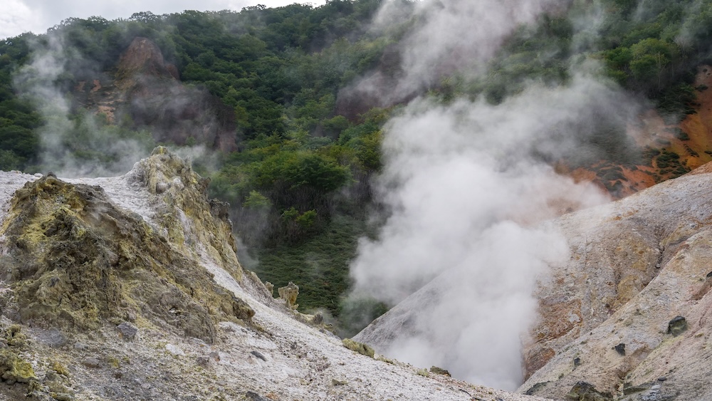 Columnas de vapor geotérmico entre rocas volcánicas de Jigokudani, Valle del Infierno, Noboribetsu, Japón Densas columnas de vapor geotérmico blanco se elevan entre formaciones de roca volcánica clara, teñidas por minerales, en Jigokudani, Valle del Infierno, Noboribetsu Onsen, Hokkaido, Japón, con laderas cubiertas de bosque verde al fondo.