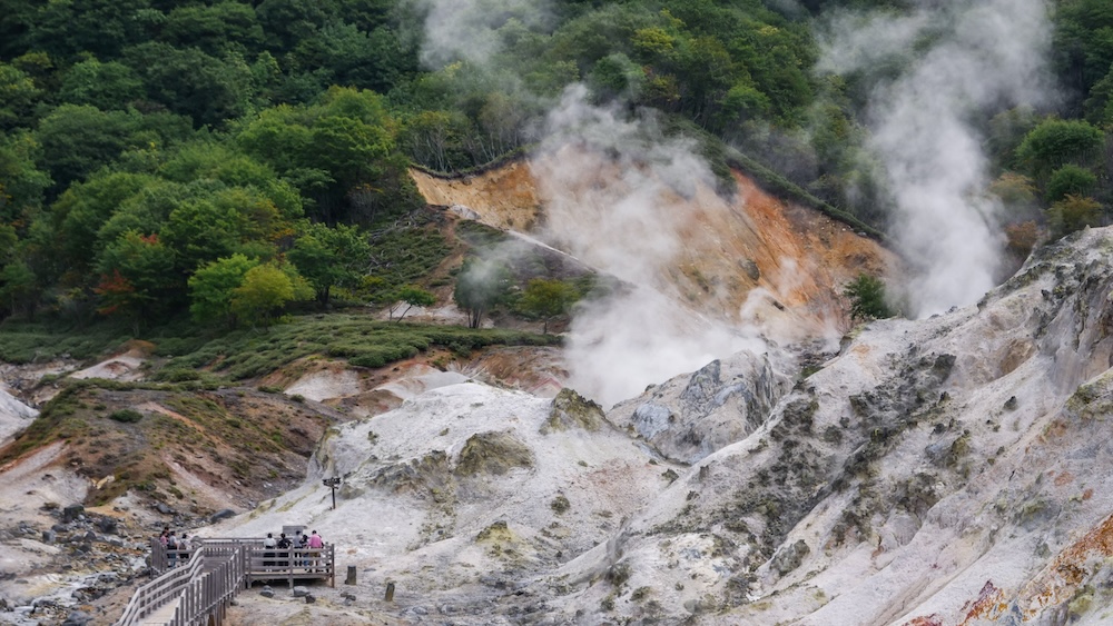 Visitantes sobre una pasarela de madera frente a rocas volcánicas blancas y naranjas y fumarolas que suben, con bosque verde de fondo, en Jigokudani, el Valle del Infierno, en Noboribetsu Onsen, Hokkaido, Japón.