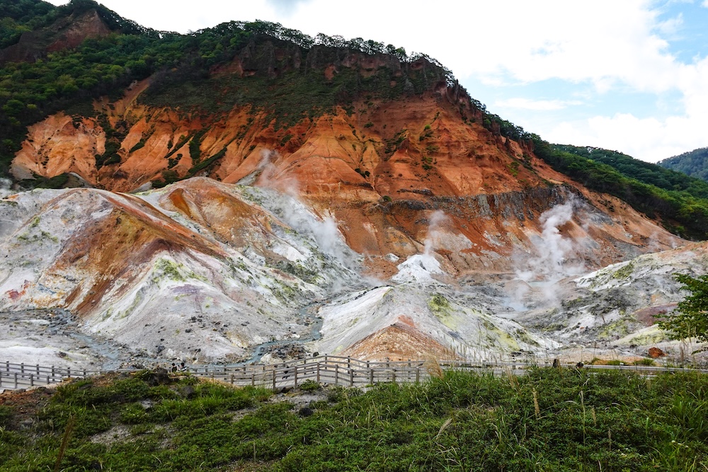 Jigokudani Hell Valley in Noboribetsu Onsen, Hokkaido, Japan – Steam Vents, Orange Cliffs and Walking Trails Wide view of Jigokudani Hell Valley in Noboribetsu Onsen, Hokkaido, Japan, showing white steam rising from mineral-stained volcanic terrain beneath orange cliffs, with wooden visitor walkways crossing the foreground.