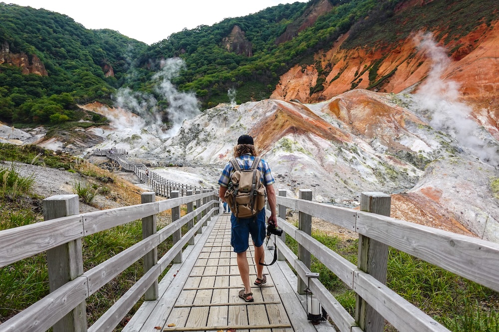 Samuel Jeffery walking along a wooden boardwalk with a backpack and camera at Jigokudani Hell Valley in Noboribetsu Onsen, Hokkaido, Japan, surrounded by steaming volcanic slopes and green forested hills.