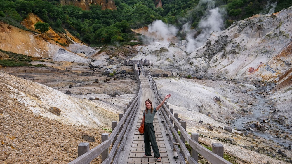 Audrey Bergner on Jigokudani Hell Valley Boardwalk Amid Steaming Volcanic Slopes – Noboribetsu, Hokkaido, Japan Audrey Bergner standing with one arm raised on a wooden boardwalk through Jigokudani Hell Valley in Noboribetsu Onsen, Hokkaido, Japan, surrounded by pale volcanic slopes, rising steam vents and green forest.
