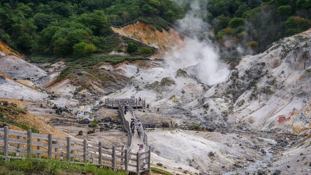 Pasarela de madera entre rocas volcánicas y vapor en Jigokudani, Valle del Infierno, Noboribetsu Onsen, Hokkaido, Japón Visitantes caminando por una pasarela de madera en Jigokudani, el Valle del Infierno de Noboribetsu Onsen, Hokkaido, Japón, rodeados de roca volcánica clara, laderas teñidas de minerales anaranjados y grandes columnas de vapor geotérmico.