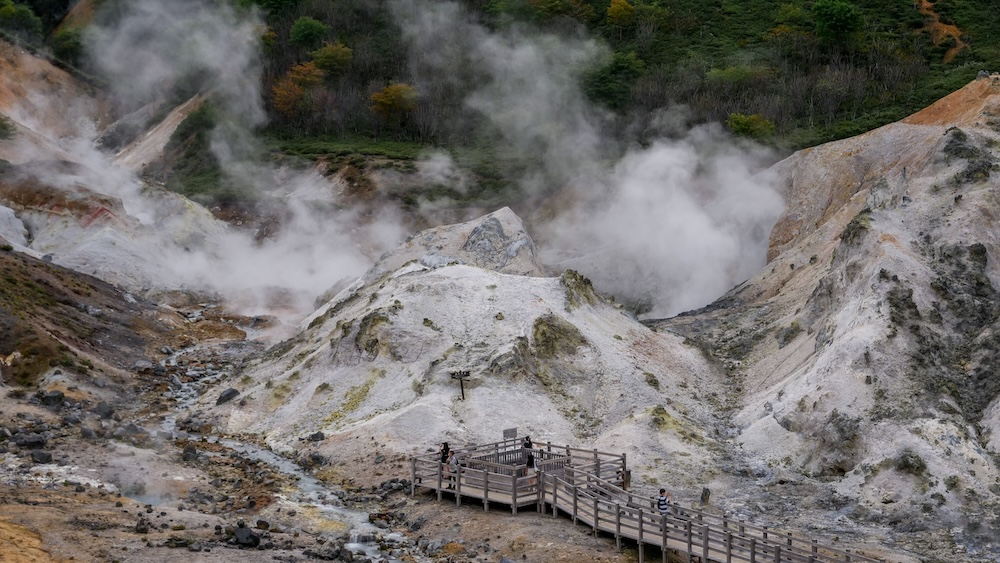 Jigokudani Hell Valley Observation Deck Surrounded by Geothermal Steam and Volcanic Slopes – Noboribetsu, Japan Wooden observation deck with several visitors at Jigokudani Hell Valley in Noboribetsu Onsen, Hokkaido, Japan, surrounded by pale volcanic slopes, rocky channels and thick plumes of geothermal steam rising toward the forest.