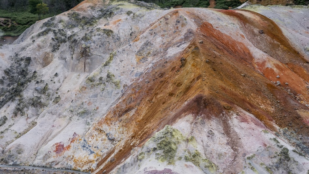 A close-up of a volcanic mound in Jigokudani (Hell Valley) shows bleached white rock streaked with rust, orange, red and pink mineral bands and yellow-green patches in Noboribetsu Onsen, Hokkaido, Japan.