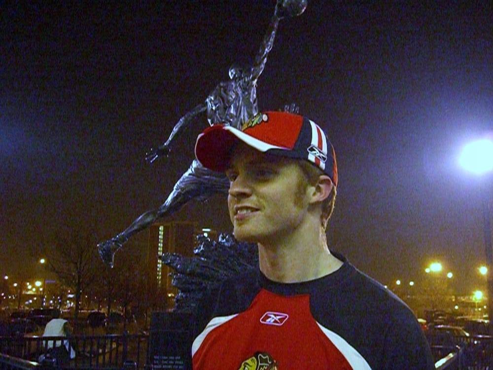 Samuel Jeffery, in his late twenties, wears a red Chicago Blackhawks cap and jersey at night beside the silhouette of the Michael Jordan statue outside the United Center in Chicago, Illinois.
