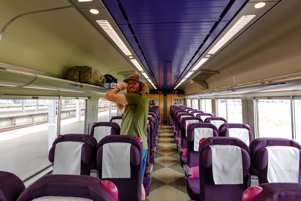 Samuel Jeffery acomodando equipaje en un tren JR de Hokkaido durante el viaje hacia Noboribetsu Onsen, Japón Samuel Jeffery acomoda el equipaje en el portavalijas superior de un tren japonés en Hokkaido, Japón, con filas de asientos tapizados de violeta, ventanas grandes y el andén de una estación visible desde el interior.