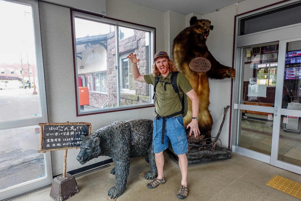 Entrada de la estación JR Noboribetsu con exhibiciones de osos pardos, Noboribetsu, Hokkaido, Japón – Samuel Jeffery Samuel Jeffery hace una pose imitando un rugido con una mochila junto a un oso pardo embalsamado y una estatua de oso en la entrada de la estación JR Noboribetsu en Noboribetsu, Hokkaido, Japón.