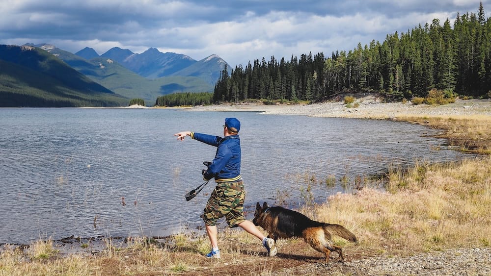 Samuel Jeffery standing beside Togo on the shore of a mountain lake in Kananaskis Country, Alberta, Canada, with evergreen forest, rocky peaks and dark clouds across the water.