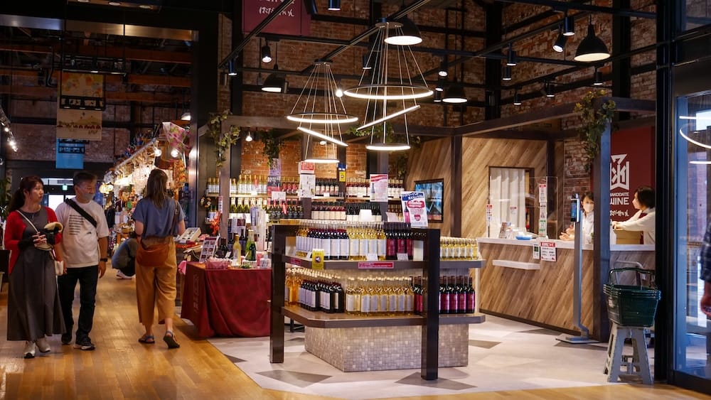 Shoppers, including Audrey Bergner, browse a brick-walled shop inside the Kanemori Red Brick Warehouses in Hakodate, Hokkaido, Japan, with wooden floors, pendant lights, wine bottle displays and staff at a counter.
