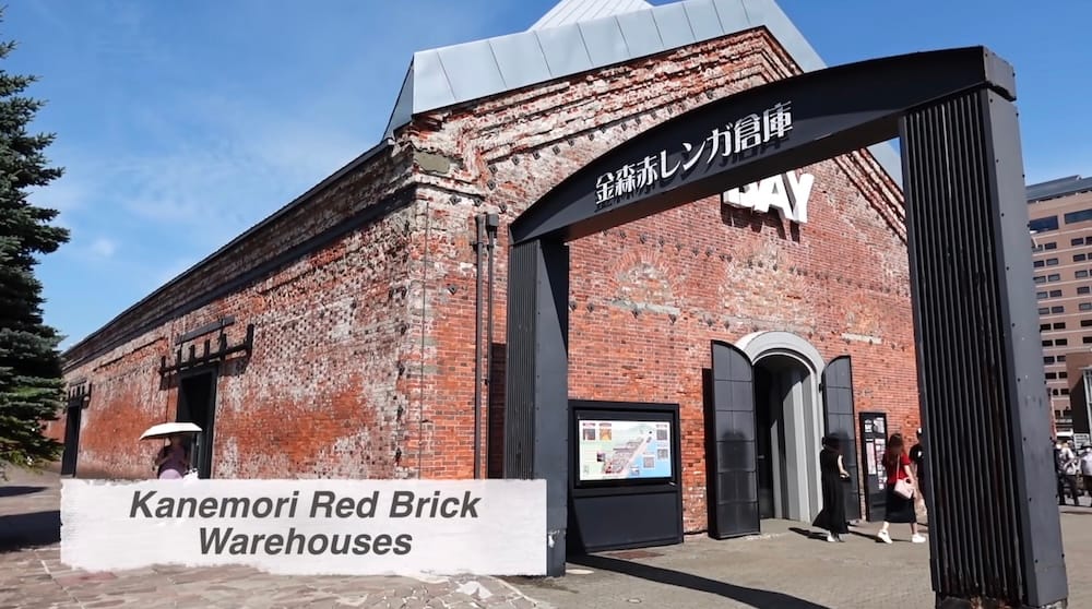 The weathered red brick front of the Kanemori Red Brick Warehouses in Hakodate, Hokkaido, Japan, with a black entrance arch bearing Japanese lettering, a site map board and visitors, one under a parasol, on a sunny day.