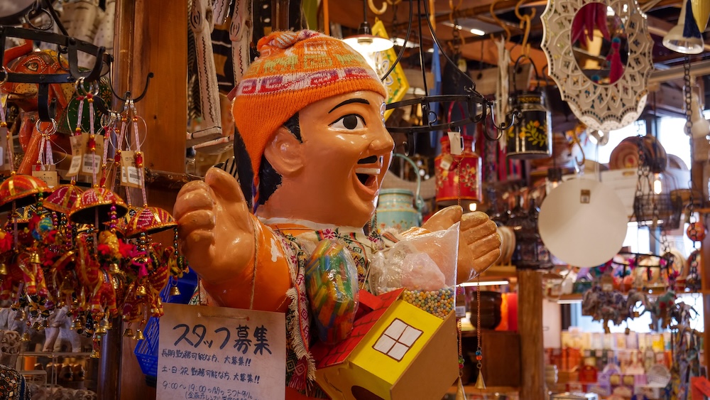 Colourful Souvenir Shop With Smiling Wooden Figurine and Hanging Crafts at Kanemori Red Brick Warehouses, Hakodate, Japan Colourful souvenir shop inside the Kanemori Red Brick Warehouses in Hakodate, Hokkaido, Japan, featuring a large smiling wooden figurine wearing an orange knitted hat, surrounded by hanging ornaments and decorative crafts.
